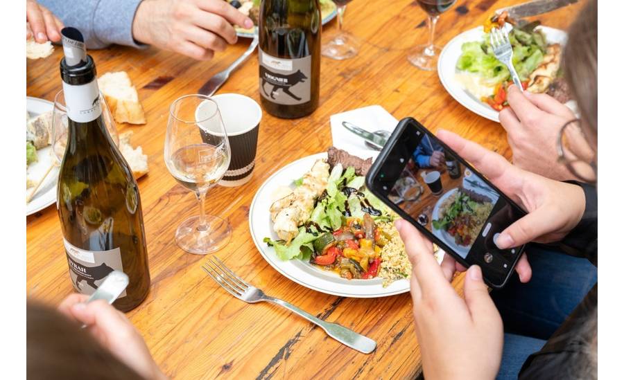 Bouteilles de vin et assiettes du brunch posées sur une table. Une personne est en train de prendre une photo avec un téléphone.
