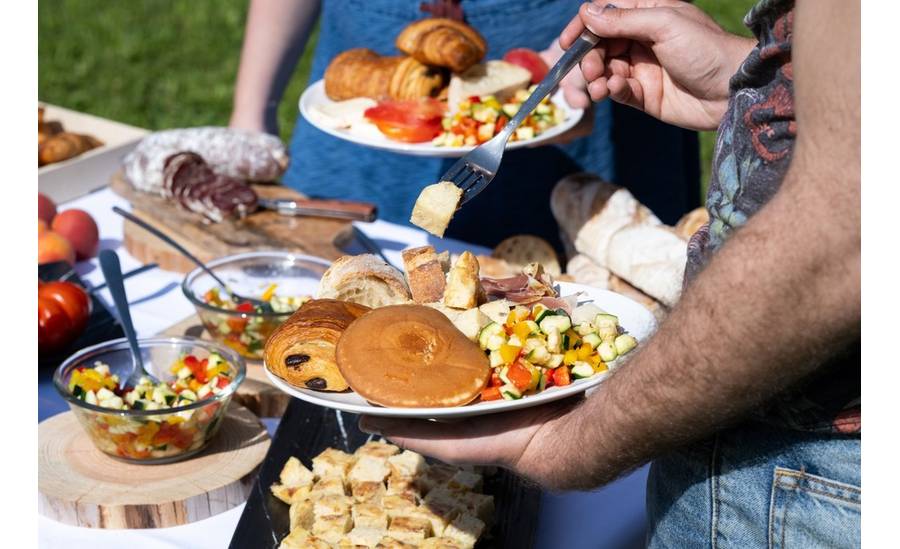 Brunch Vigneron installé sur une table avec une nappe blanche posée au milieu de l'herbe. Une personne est en train d'attraper de la nourriture avec une fourchette.
